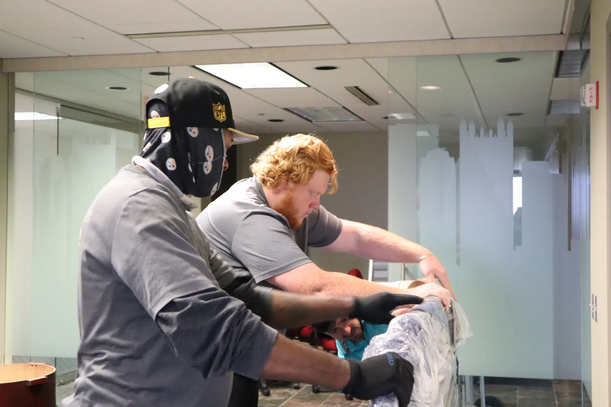 Movers handling a large wrapped item inside a glass-walled office.