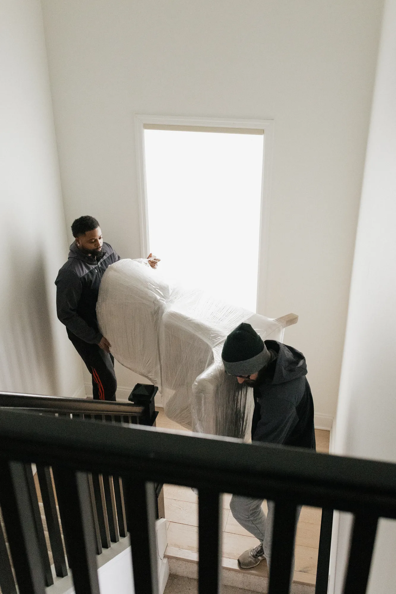 Two movers carrying plastic-wrapped furniture down an indoor staircase.