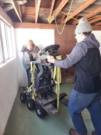 Two movers moving a heavy strapped workshop machine on a dolly.