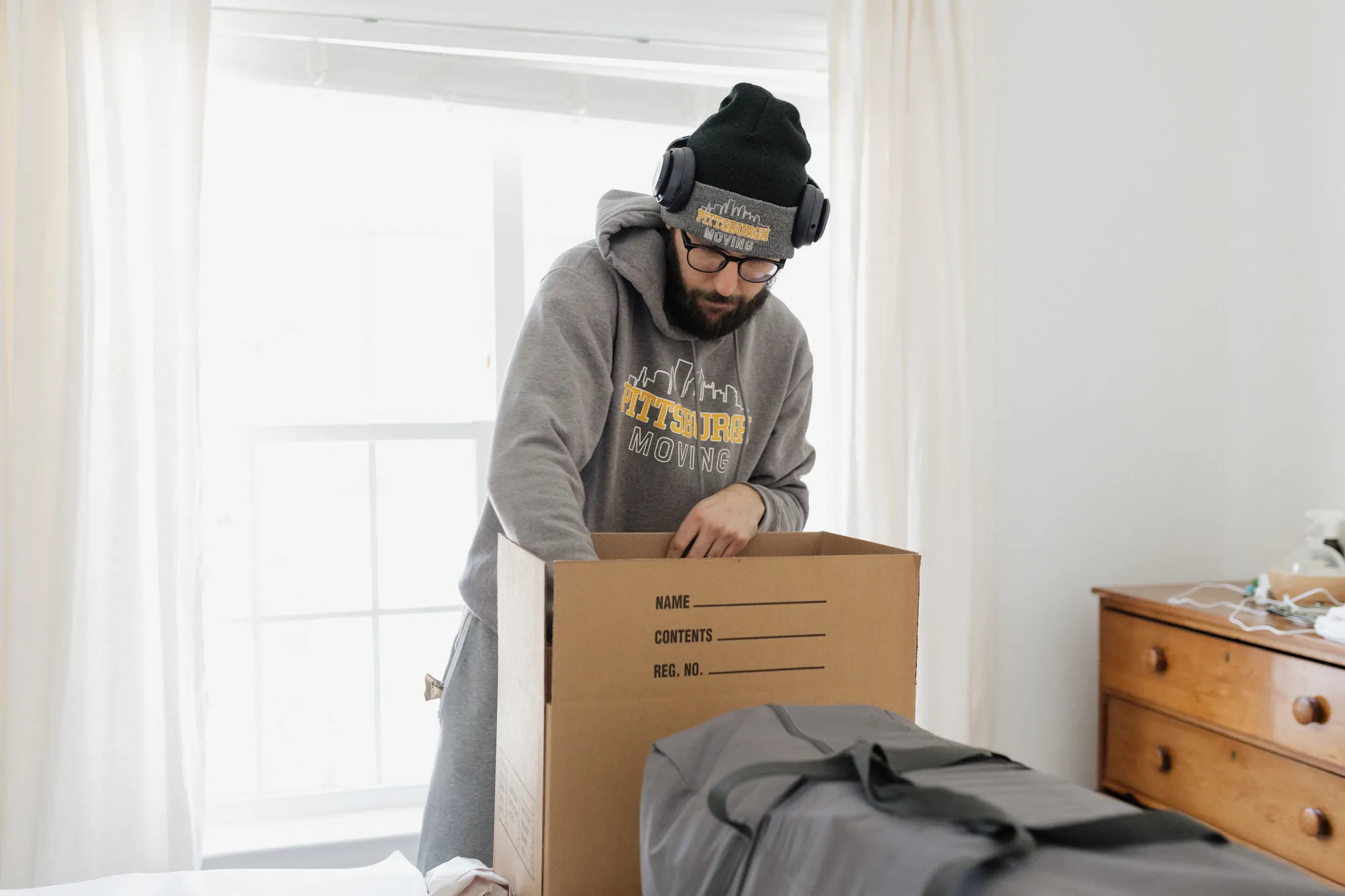 Pittsburgh Moving PGH packer in a company hoodie filling a moving box beside a dresser.