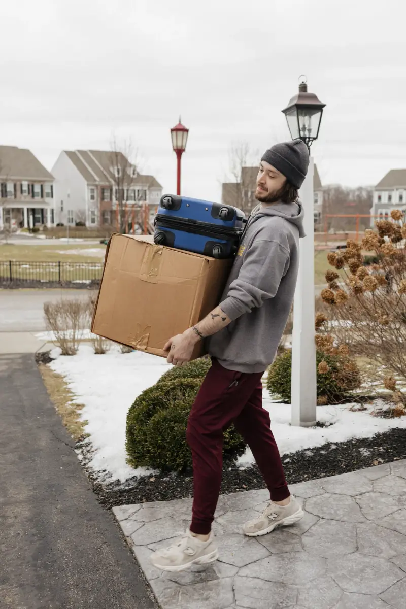 Mover carrying a large moving box and a suitcase down a snowy driveway.