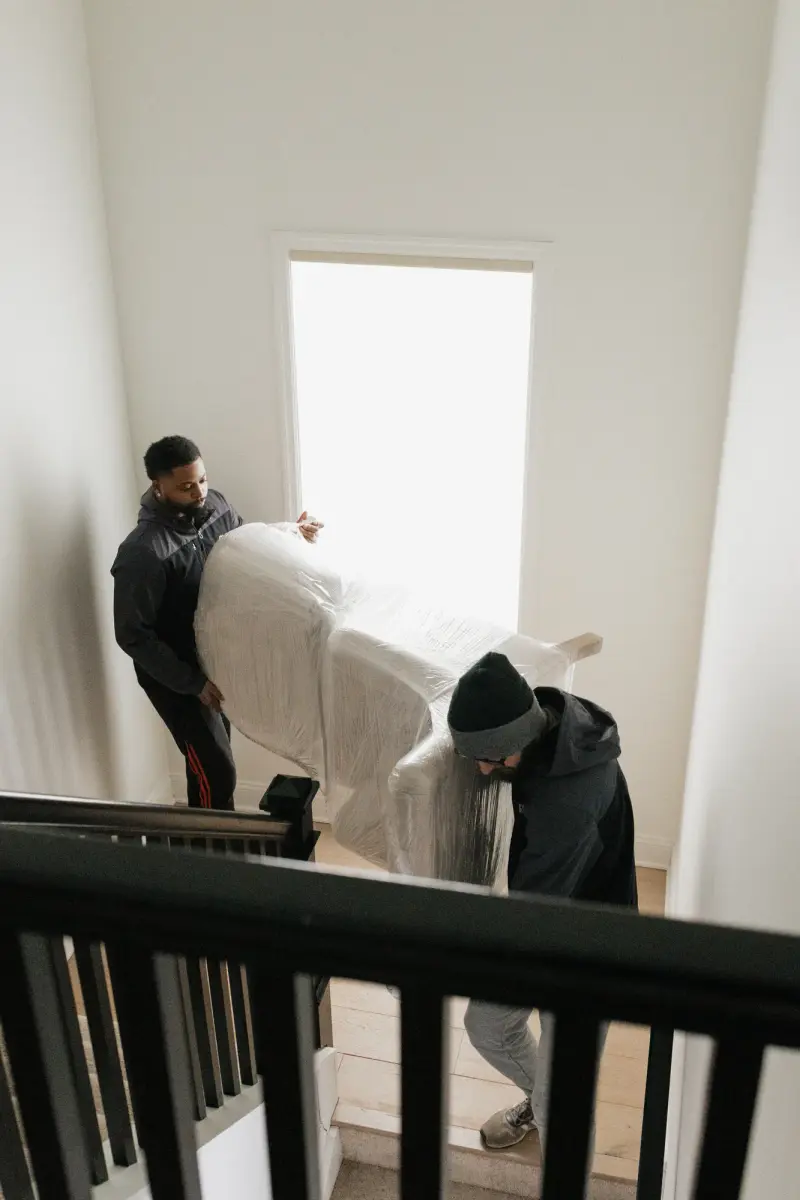 Two movers carrying plastic-wrapped furniture down an indoor staircase.