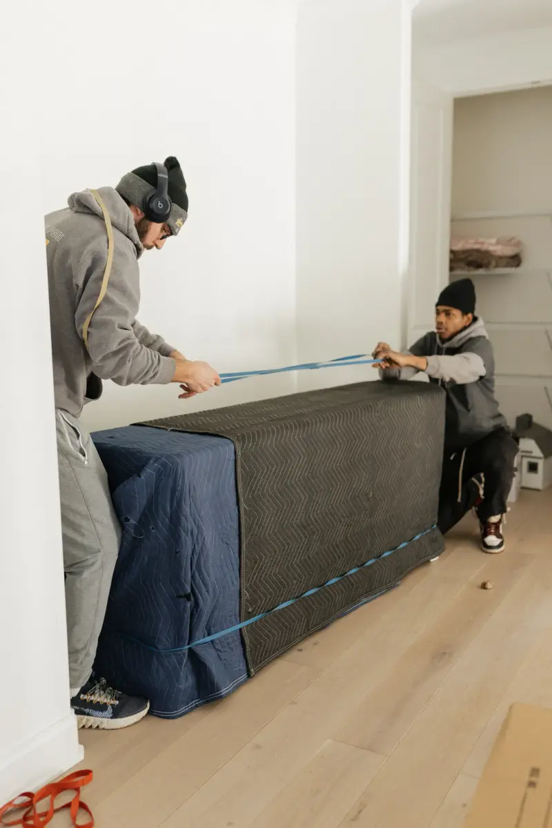 Two movers strapping a large blanket-wrapped item before carrying it out of a home.