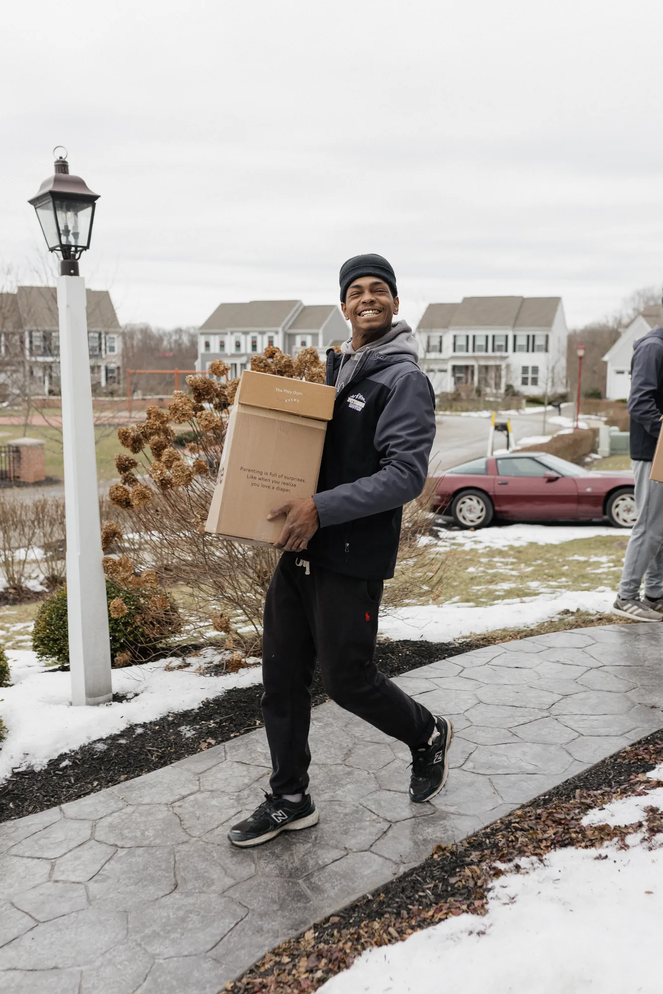 Smiling mover carrying a moving box along a snowy walkway during a winter move.